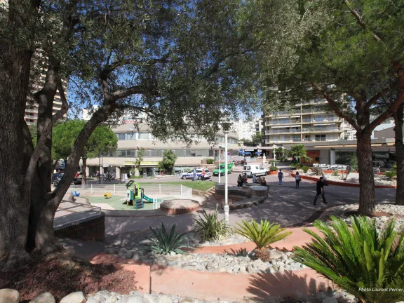 Chemin entouré d'arbustes et de galet menant à une place avec en son centre une aire de jeux pour enfants
