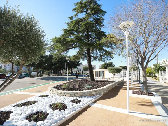 Parterre de galets et de plantes entouré d'un large trottoir, d'arbres et de lampadaires.