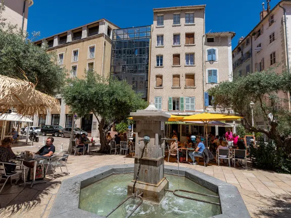 AU centre de la place, fontaine en pierre. Autour, terrasses de restaurant.