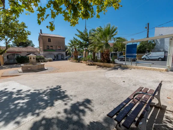 Vue sur la place donnant sur une pharmacie. Un banc à l'ombre d'un arbre fait face à une fontaine et à la droite de la place se trouve un arrêt de bus.