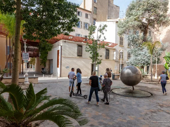Vue sur la place végétalisée sur laquelle des passants déambulent avec au fond vue sur la maison de la photographie et au centre le fameux globe terrestre.