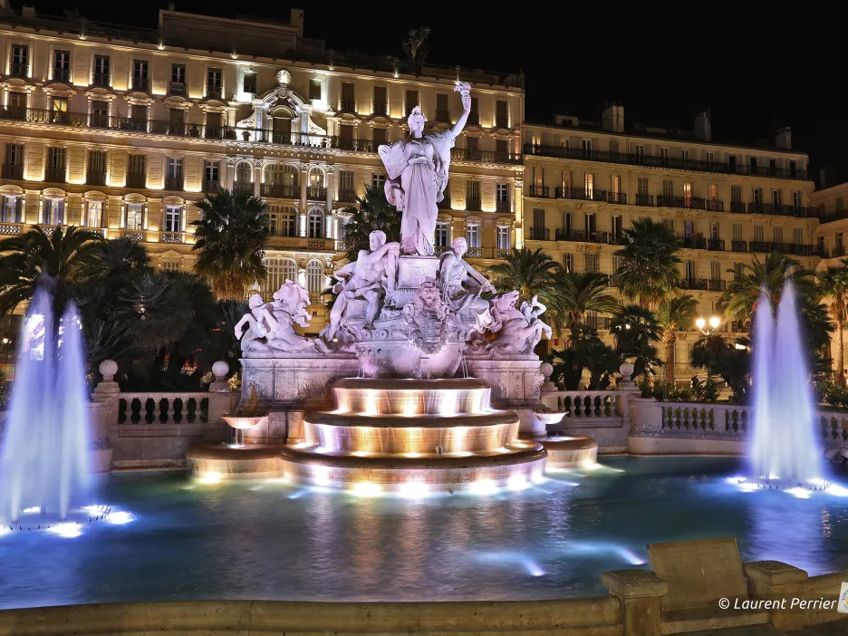 Photographie de nuit de la place de la Liberté : au centre, la fontaine et sa statue, ornée de luminaires éclairant l'ensemble des éléments de la fontaine. Derrière la fontaine, un bâtiment également éclairé de toutes parts