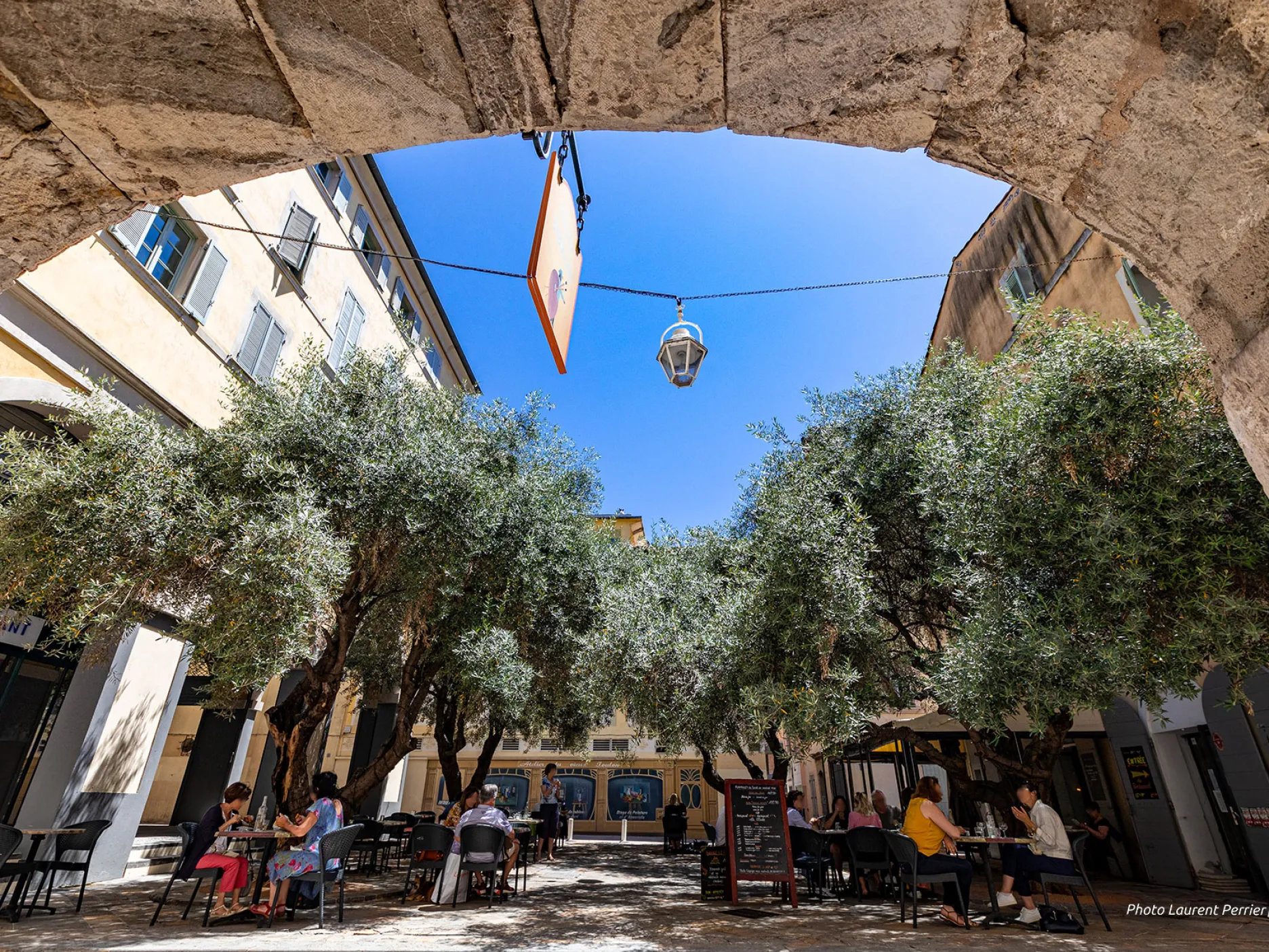 Vue sur la place ombragée grâce à des oliviers et remplie de tables et de chaises d'un restaurant