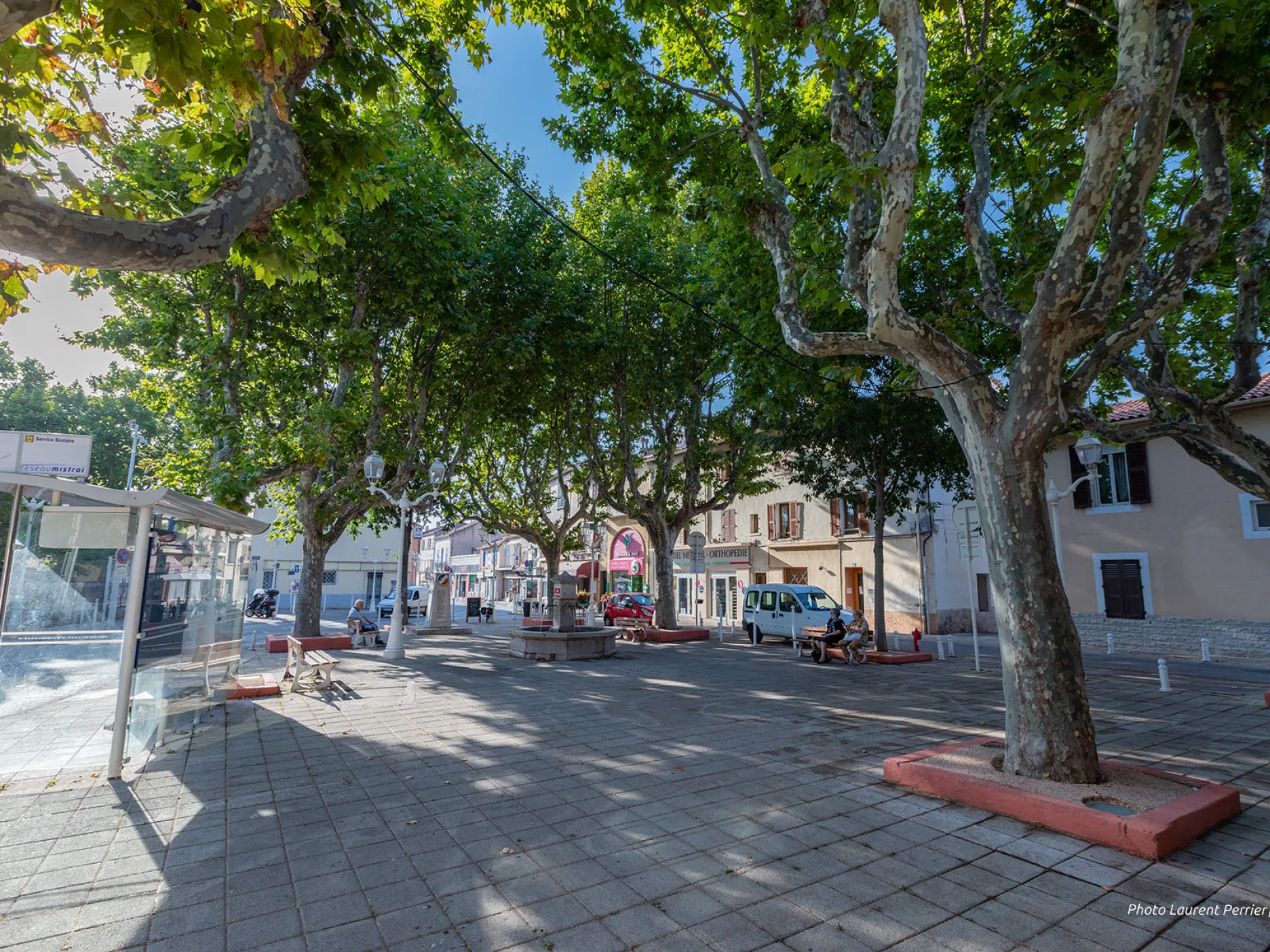 Place dallée avec des arbres, des bancs ainsi qu'une fontaine.