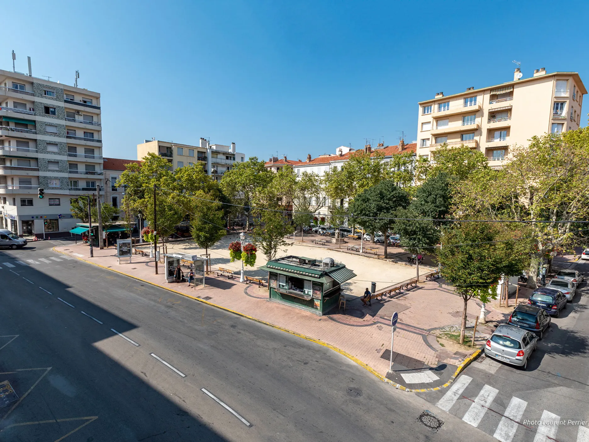 Vue aérienne du boulodrome de la place ainsi qu'un kiosque. Ceux ci sont logés par des voitures garés sur le bas côté et entourés par des arbres.