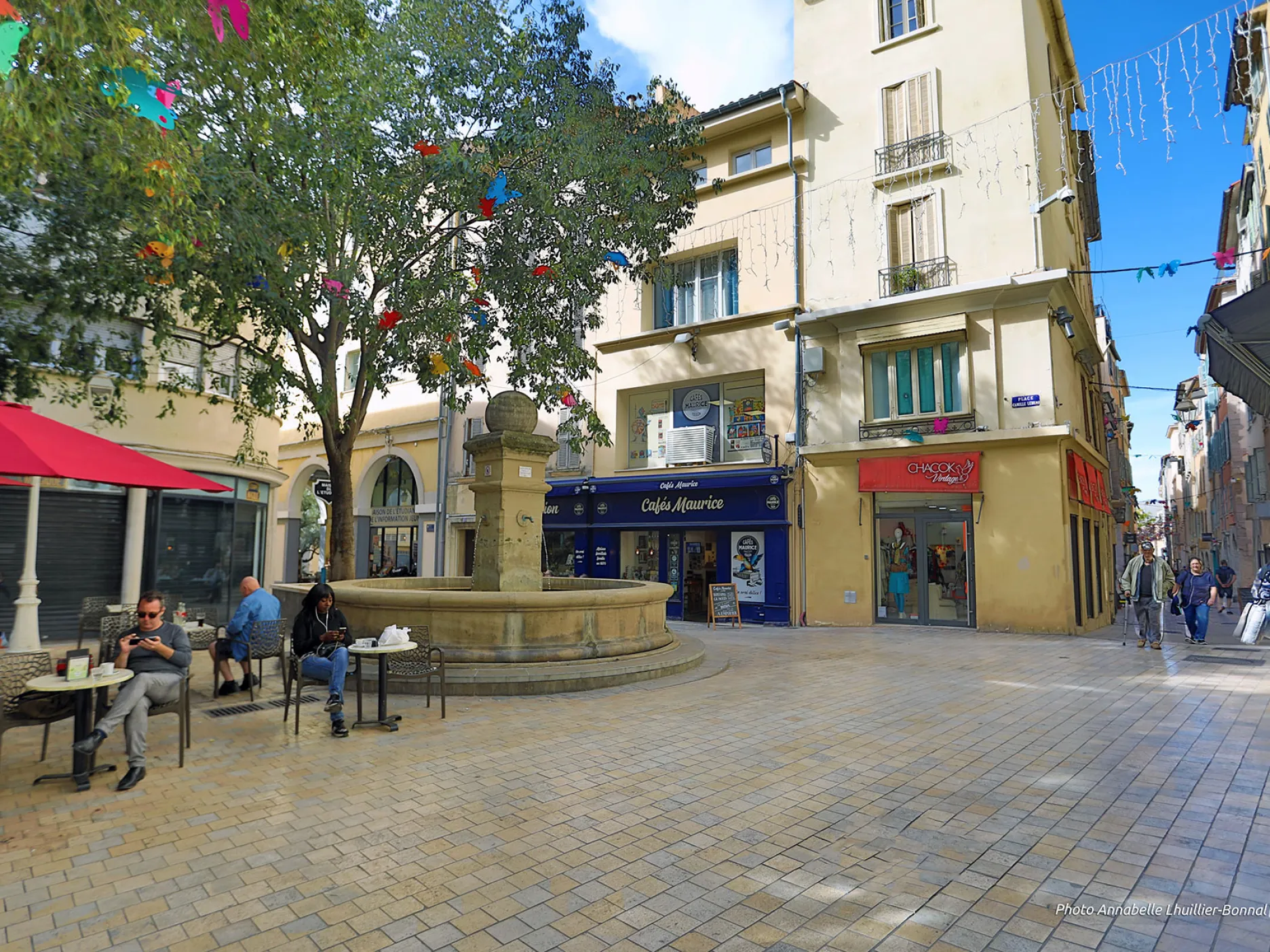 Vue sur une rue piétonne longeant la place couverte d'une terrasse de café collée à une fontaine.