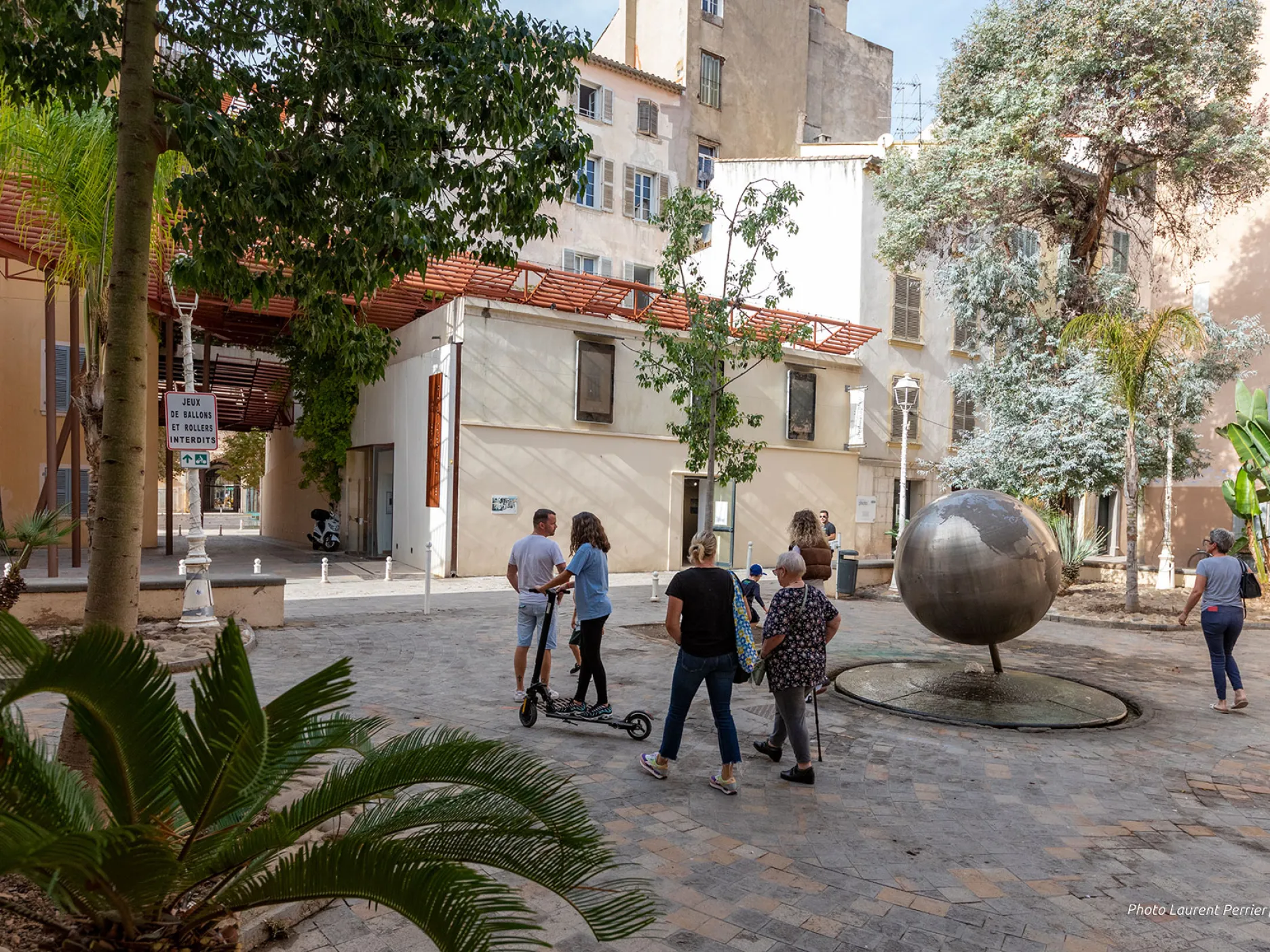 Vue sur la place végétalisée sur laquelle des passants déambulent avec au fond vue sur la maison de la photographie et au centre le fameux globe terrestre.